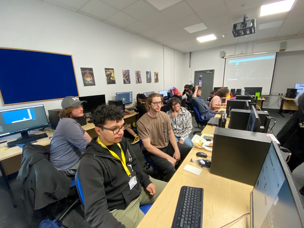 An image of the first UWL game jam in 2021. Students are sat in a classroom and looking at a computer screen. A student in the middle is holding a thumbs up gesture, while another has both hands on their head.
