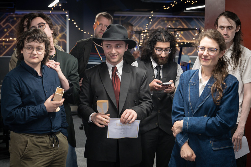A group of students wearing smart clothes are stood together, some holding small golden awards with the UWL logo on them. The picture is taken in a computing room with upright PC towers with LED lights and gaming chairs, and fairy lights are draped from the ceiling in the background.