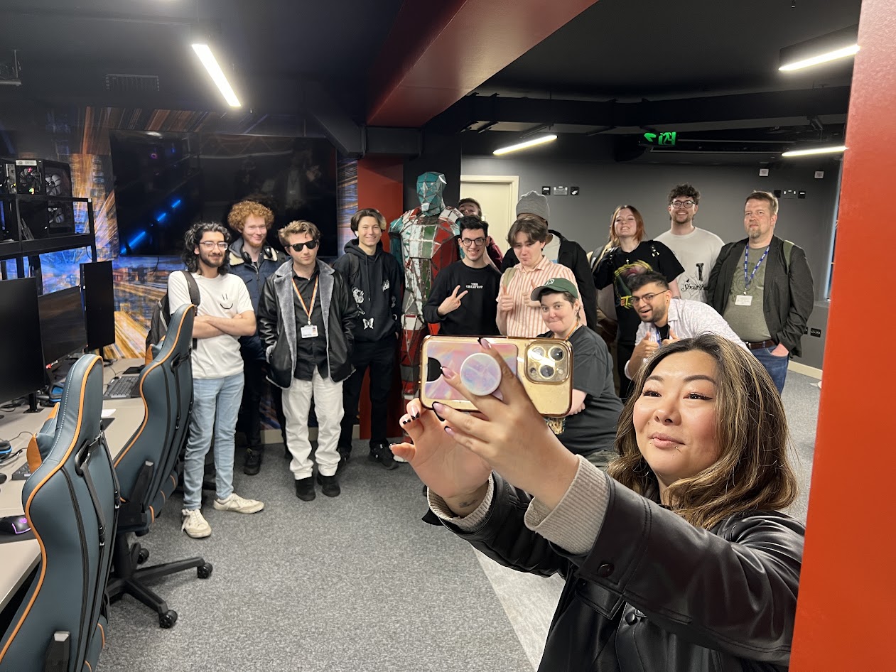 An image of the first UWL game jam to take place in the Digital Futures Lab in 2024. A lecturer is holding a phone in selfie mode to take a picture with students and staff in a high-tech looking room with dual-monitor PCs and gaming chairs.