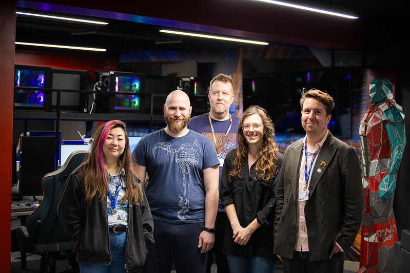 An image of 5 people standing in a computing room facing the camera. Behind them are computer towers with bright LEDs and gaming chairs. There are 3 UWL lecturers and 2 visitors from SEGA Europe.